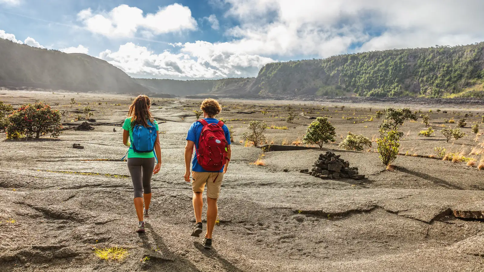 Two hikers crossing the flat solidified lava lake on the floor of Kīlauea Iki crater, with the crater walls rising behind them