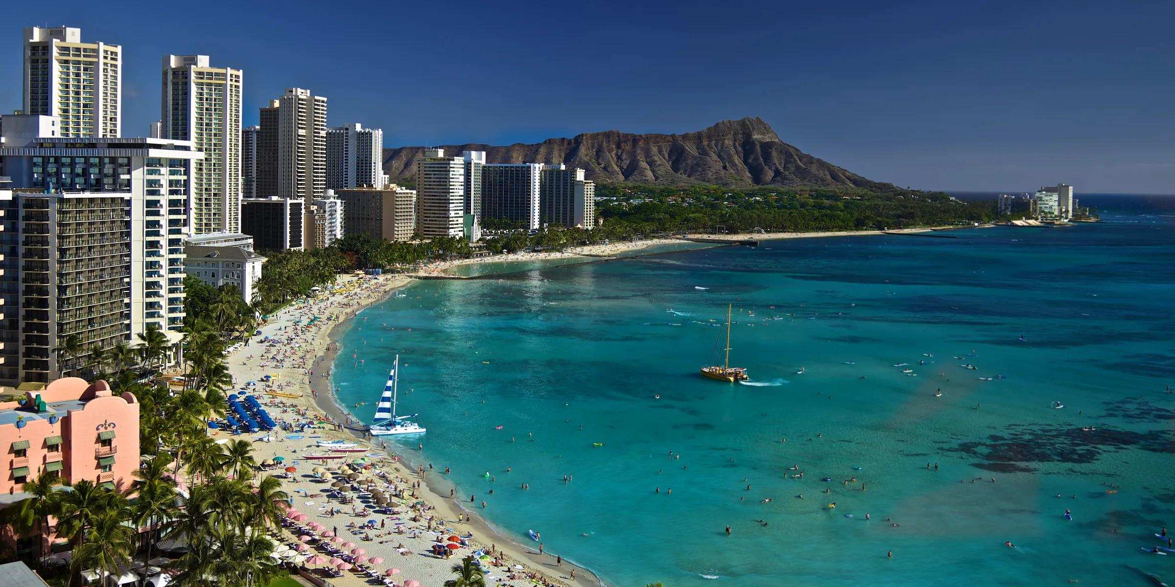 Busy Waikiki Beach with a sailing catamaran offshore and Diamond Head behind