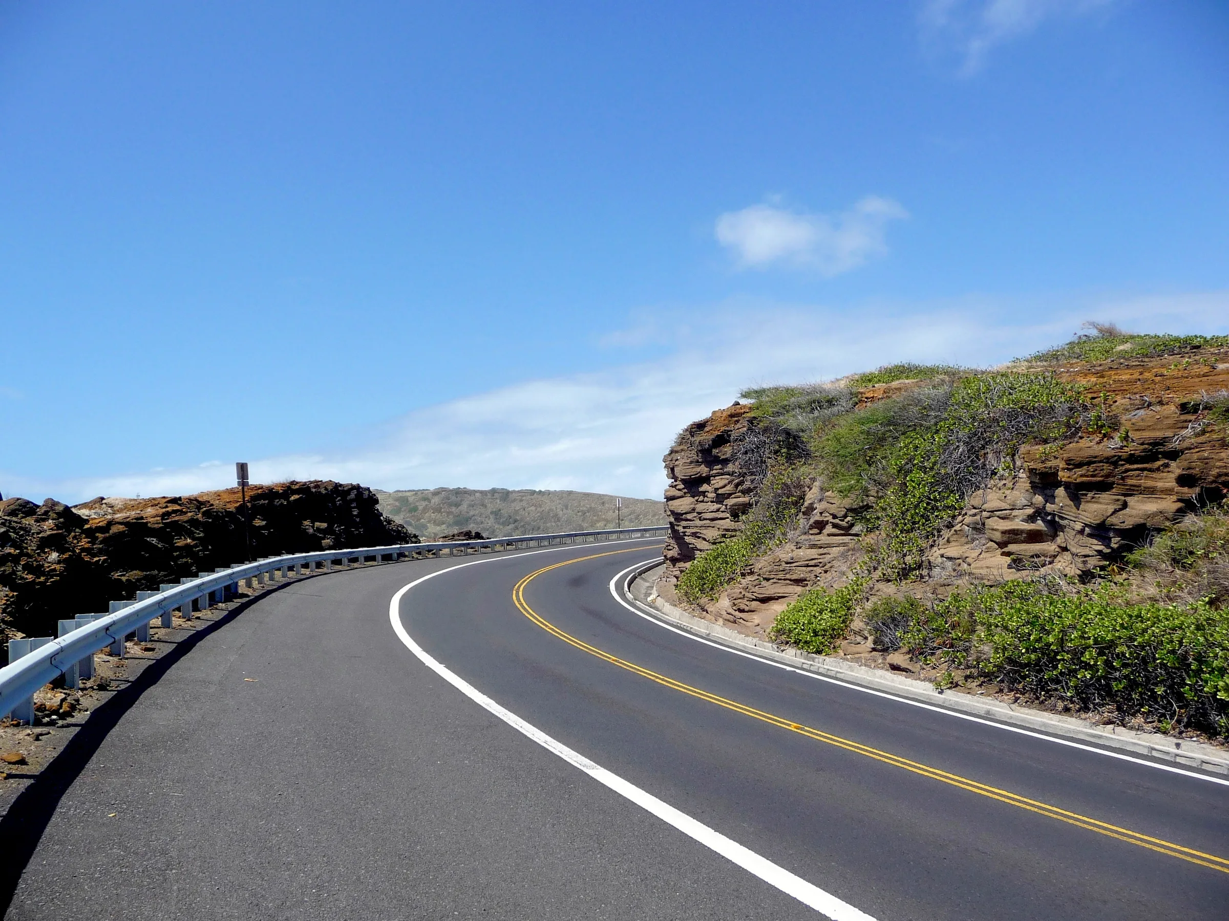 Two-lane coastal road curving past rocky cliffs under a blue sky